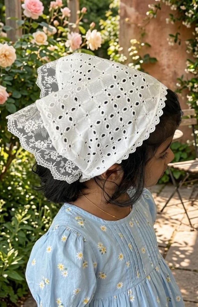 May include: A white eyelet bonnet with a scalloped lace trim. The bonnet is worn over dark hair, paired with a light blue dress with a daisy pattern. The background features a garden with pink roses and greenery.