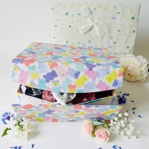 May include: A gift box with a colorful gummy bear pattern, open to reveal contents. A matching gift bag with a lace bow sits behind the box. Flowers, including roses and blue cornflowers, are arranged around the box, creating a cheerful presentation.