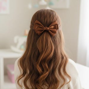 Puede incluir: Un lazo de satén marrón para el cabello es el punto focal de esta imagen. El lazo está centrado en el cabello, que es largo y ondulado. El cabello es de un color marrón cálido. El fondo es suave y desenfocado.