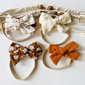 May include: Four fabric baby headbands with bow accents. The bows are in cream, rust, and brown colors, with floral and textured patterns. The headbands are on a neutral background with a macrame wall hanging.