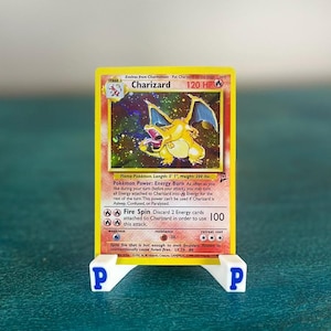 May include: A Charizard Pokémon card, displayed on a white stand with blue "P" letters. The card is yellow with a holographic image of a dragon-like creature. Text includes "Charizard" and "120 HP".