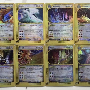 May include: Eight collectible Pokémon trading cards featuring various characters like Charizard, Lugia, and Nidoking. Each card has a gold border, character illustrations, and text detailing abilities and HP. The cards are displayed on a light-colored surface.