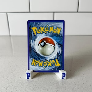 May include: A Pok&eacute;mon trading card with a blue background and the Pok&eacute;mon logo in yellow. The card features a red and white Pok&eacute; Ball design. The card is displayed on a white stand with the letter "P" on each side.