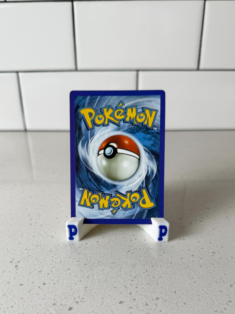May include: A Pok&eacute;mon trading card with a blue background and the Pok&eacute;mon logo in yellow. The card features a red and white Pok&eacute; Ball design. The card is displayed on a white stand with the letter "P" on each side.