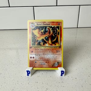 May include: A yellow Blaine's Charizard Pokémon card, featuring a fiery orange dragon illustration. The card is displayed on a white stand with blue "P" letters. The card's text includes "100 HP" and "Flame Jet".