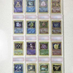 May include: A collection of sixteen Pokemon trading cards, each encased in a protective plastic sleeve. The cards feature various colorful illustrations of Pokemon characters, with text and symbols. The cards are arranged in four rows of four.