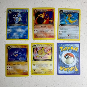 Może przedstawiać: Sześć kart kolekcjonerskich Pokémon. Karty przedstawiają różne postacie, w tym Dark Charizard, Dark Blastoise i Dark Dragonite. Karty są żółte i niebieskie z ilustracjami postaci i tekstem.