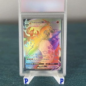 Puede incluir: Una carta Pokémon Charizard VMAX graduada, con un diseño holográfico de arcoíris. La carta está dentro de un protector transparente y se muestra en un soporte blanco. El texto de la carta incluye "Claw Slash" y "G-Max Wildfire".