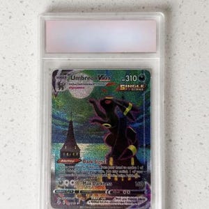 May include: A holographic Umbreon VMAX Pokémon card in a protective case. The card features a dark purple and black Umbreon illustration with the text "Umbreon VMAX" and "Dark Signal". The card is graded and encased in a clear plastic holder.