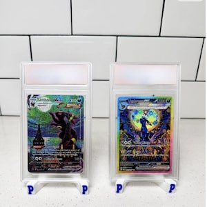Puede incluir: Dos cartas coleccionables de Pokémon en fundas protectoras. Las cartas presentan ilustraciones de Umbreon con colores vibrantes y texto. Cada carta se muestra en un soporte blanco con un logotipo "P" azul. El fondo es una pared de azulejos blancos.