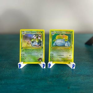 May include: Two Pokémon trading cards, "Erika's Venusaur" and "Venusaur", are displayed on white stands with the letter "P". The cards have yellow borders and feature illustrations of the Pokémon. The "Erika's Venusaur" card shows 90 HP, while the "Venusaur" card shows 100 HP.