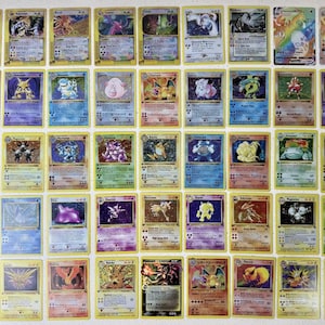 Puede incluir: Una colección de cartas coleccionables Pokémon vintage, con varios personajes y diseños. Las cartas tienen bordes amarillos y están dispuestas sobre una superficie de color claro. Las cartas muestran una variedad de colores e ilustraciones.