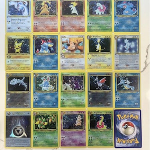 Puede incluir: Una colección de cartas coleccionables de Pokémon, con varios personajes con ilustraciones coloridas y texto. Las cartas tienen bordes amarillos y están dispuestas sobre una superficie clara. La imagen incluye una carta con el logotipo de Pokémon.