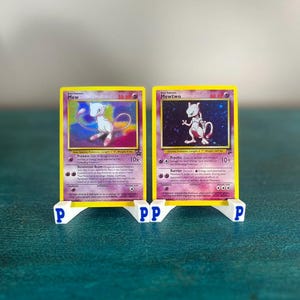 May include: Two Pokémon trading cards, "Mew" and "Mewtwo", displayed in yellow and white card holders. The cards feature illustrations of the Pokémon characters. The holders are white with the letters "P" printed on them. The background is a teal color.