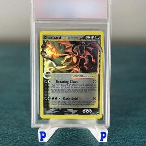 May include: A Charizard Pokémon trading card in a protective case. The card features a fiery dragon illustration with the text "Charizard" and "90 HP". The card is displayed on a blue surface with white supports.