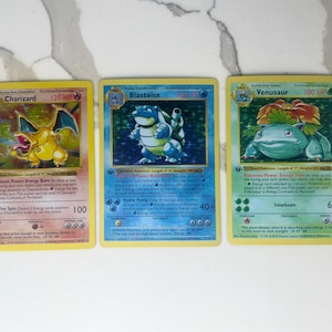 May include: Three Pokémon trading cards featuring Charizard, Blastoise, and Venusaur. The Charizard card has a yellow border and shows a yellow dragon. The Blastoise card has a blue border and shows a blue turtle. The Venusaur card has a green border and shows a green plant-like creature.
