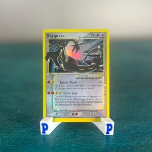 May include: A yellow Rayquaza Pokémon card with a black and red illustration, displayed on a white and blue stand. The card features text including "Rayquaza" and "90 HP". The stand has the letter "P" on each side.