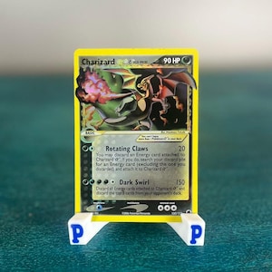 May include: A Charizard Pokémon trading card, featuring a dragon-like creature with orange and black coloring, is displayed on a white stand with blue "P" logos. The card has text that reads "Charizard" and "90 HP".
