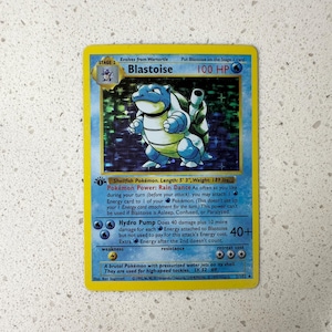 Puede incluir: Carta coleccionable de Pokémon Blastoise. Presenta una criatura azul con forma de tortuga con dos cañones en la espalda. La tarjeta tiene un borde amarillo e incluye texto sobre las habilidades y estadísticas del personaje.