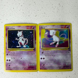 May include: Two Pokémon trading cards featuring Mewtwo and Mew. The Mewtwo card has a purple and black background with a white and purple Mewtwo illustration. The Mew card has a pink and purple background with a white and pink Mew illustration.
