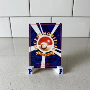 Puede incluir: Una carta coleccionable de Pok&eacute;mon, con un dise&ntilde;o de Pok&eacute; Ball rojo y blanco, se muestra en un soporte blanco con letras "P" azules. El reverso de la carta tiene un fondo azul con rayas blancas y las palabras "Pocket Monsters Card Game".