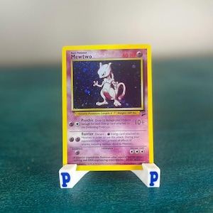 May include: A Pokémon trading card featuring Mewtwo, a Psychic-type Pokémon. The card has a yellow border and displays the character with a purple and white color scheme. The card is held by a white stand with the letter "P" on each side.