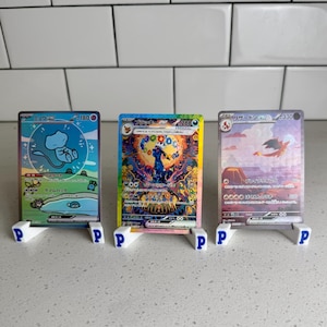 May include: Three Pokémon trading cards displayed on white stands with the letter "P". The cards feature colourful illustrations and Japanese text. The stands are on a white surface.