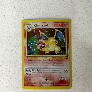 Peut inclure: Une carte Pokémon Charizard. La carte présente une illustration de Charizard jaune et orange avec des ailes bleues. Le texte de la carte comprend "Charizard", "120 HP" et "Fire Spin". C'est une carte de niveau 2.