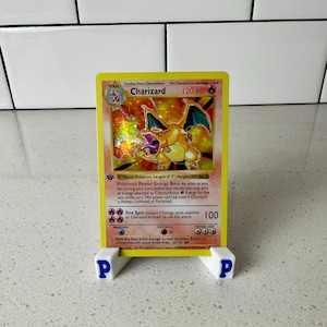 May include: A Charizard Pokémon card, featuring a fiery orange dragon, is displayed on a white stand with blue "P" logos. The card has a yellow background with text and illustrations.