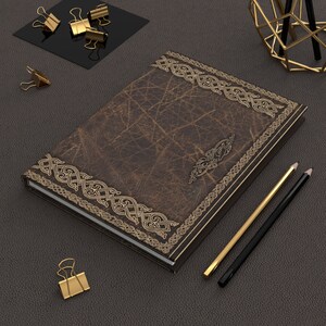 May include: A brown leather journal with gold Celtic knotwork border and accents. Gold binder clips, pencils, and a geometric pen holder are arranged around the journal on a dark gray surface. The journal has a distressed leather texture.