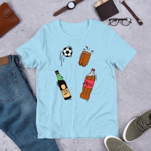 May include: Light blue t-shirt featuring a soccer ball, a brown pastry, and two bottles with text. One bottle is green with "FER-NET" on it, and the other is brown with "Vigris" on it. The shirt is made of a soft material.