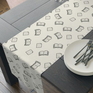 May include: A white table runner with a black and white book pattern. The runner is on a dark wood table with a bowl of apples, two wine glasses, and a plate of silverware.