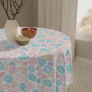 May include: A round white tablecloth with a repeating pattern of pastel clouds, rainbows, and crescent moons. The design includes pink, blue, and yellow elements. A white vase, a small basket of fruit, and two bowls are on the table.