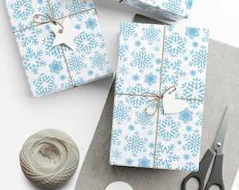 May include: Three wrapped gifts with light blue snowflake patterned paper, tied with twine and white tags. A ball of twine, scissors, and blank tags are also present, suggesting a holiday or winter theme.