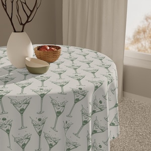 May include: A round tablecloth with a repeating pattern of green martini glasses with olives. The tablecloth is on a round table with a vase, a bowl, and a basket of fruit. The background includes a window and a curtain.