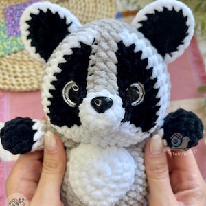 May include: A plush raccoon toy made of soft, chunky yarn in shades of grey, black, and white. The stuffed animal features large, glossy black eyes with silver accents, a small black nose, and a white belly patch. The logo CuteVilleToys is visible.