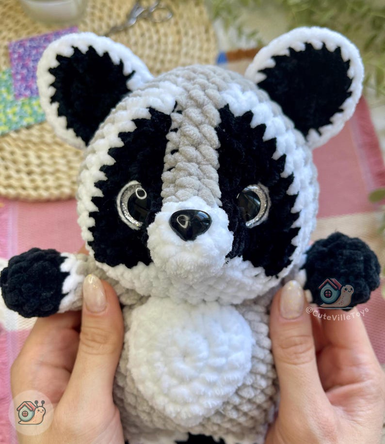 May include: A plush raccoon toy made of soft, chunky yarn in shades of grey, black, and white. The stuffed animal features large, glossy black eyes with silver accents, a small black nose, and a white belly patch. The logo CuteVilleToys is visible.