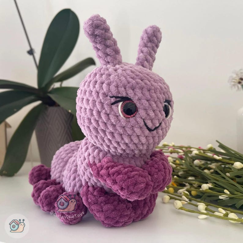 Puede incluir: Un juguete de oruga de peluche hecho a mano en tonos de morado y magenta. La oruga tiene ojos grandes y expresivos, una boca sonriente y dos antenas verticales. El juguete est&aacute; hecho de hilo suave y texturizado y est&aacute; dise&ntilde;ado para ser tierno.