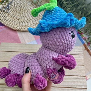 Puede incluir: Un juguete de oruga de ganchillo hecho a mano en tonos de morado, azul y verde. La oruga tiene un sombrero azul con detalles verdes y pies morados. El juguete es suave y afelpado, ideal para ni&ntilde;os.