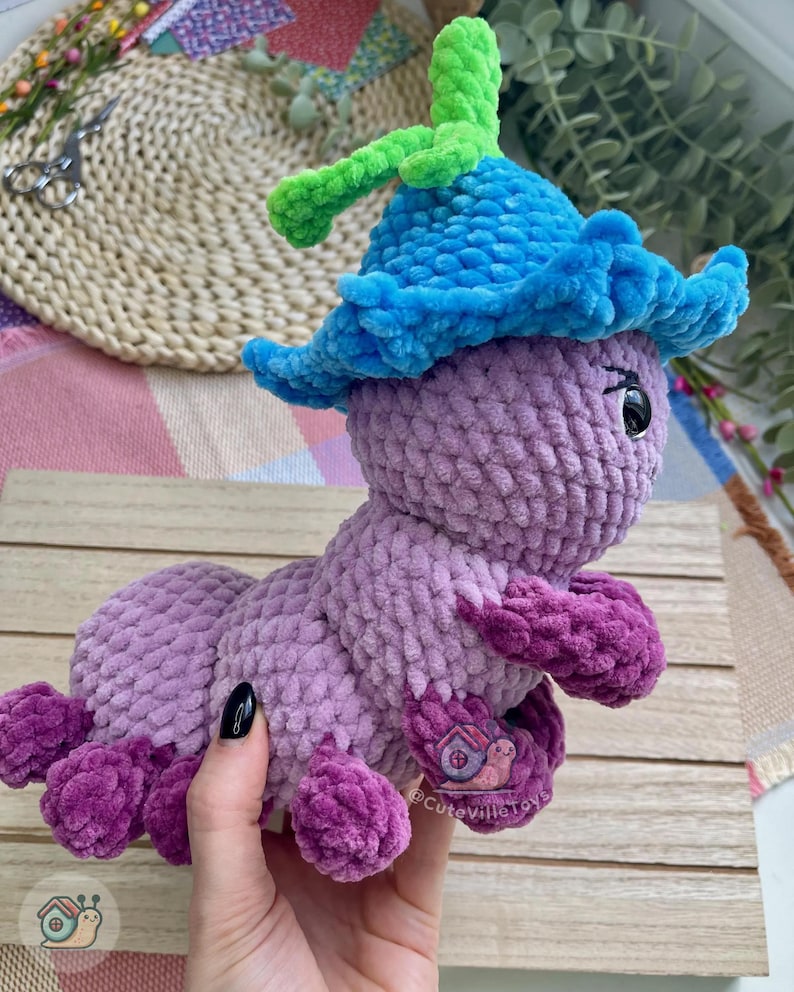 Puede incluir: Un juguete de oruga de ganchillo hecho a mano en tonos de morado, azul y verde. La oruga tiene un sombrero azul con detalles verdes y pies morados. El juguete es suave y afelpado, ideal para ni&ntilde;os.