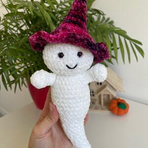Op de afbeelding: Witte gehaakte geest knuffel met een roze en paarse fluwelen heksenhoed met zwarte accenten. De geest heeft zwarte ogen en een glimlachende mond.