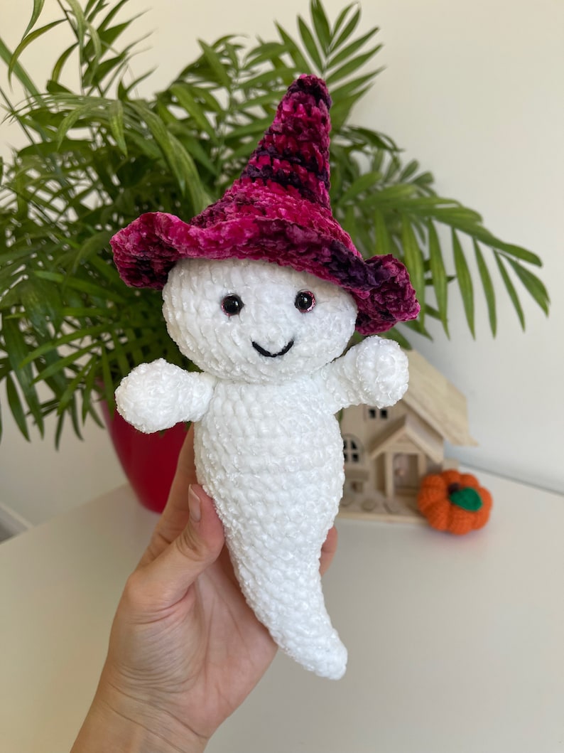 Op de afbeelding: Witte gehaakte geest knuffel met een roze en paarse fluwelen heksenhoed met zwarte accenten. De geest heeft zwarte ogen en een glimlachende mond.