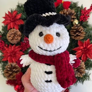 May include: A white crocheted snowman with a black velvet top hat, a red velvet scarf, and black eyes and buttons. The snowman is holding a red velvet scarf in its hands.