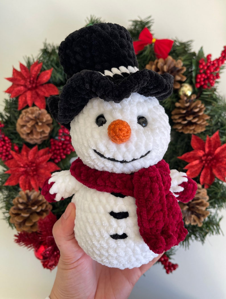 May include: A white crocheted snowman with a black velvet top hat, a red velvet scarf, and black eyes and buttons. The snowman is holding a red velvet scarf in its hands.