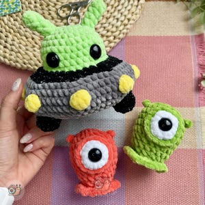 May include: A crochet UFO toy with a light green top, a dark gray base, and yellow circular accents, accompanied by two small, one-eyed crochet monster figures in bright orange and light green, all crafted from soft, plush yarn.