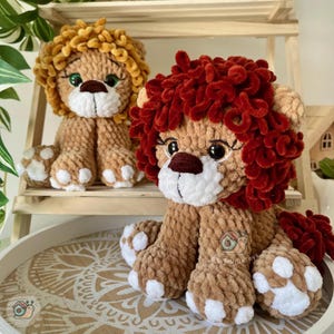 Puede incluir: Dos leones de peluche hechos de hilo suave y texturizado. Un león tiene una melena de color amarillo dorado, mientras que el otro presenta una melena de color rojo intenso. Ambos tienen cuerpos color canela, hocicos blancos y ojos grandes.