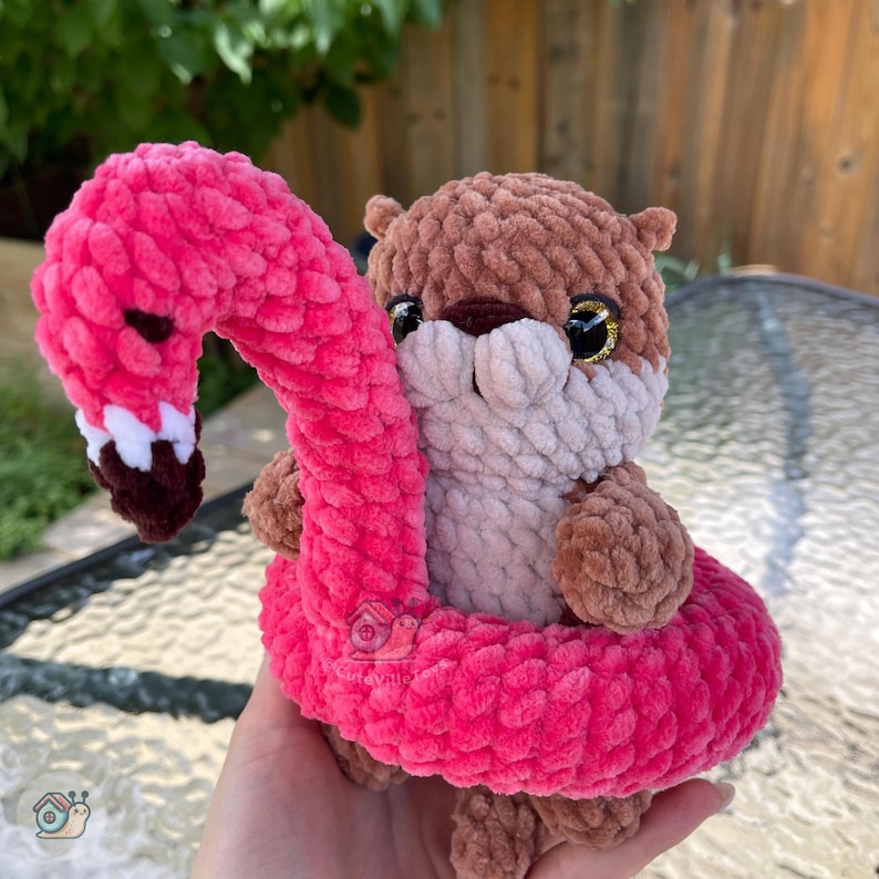 May include: A brown and light beige crocheted otter plush toy sitting inside a bright pink crocheted flamingo-shaped pool float. The otter has dark brown eyes and a soft, textured yarn finish. The flamingo float features a long neck and head.