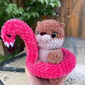 May include: A brown and light beige crocheted otter plush toy sitting inside a bright pink crocheted flamingo-shaped pool float. The otter has large, shiny black eyes with gold accents, and the flamingo float features a long, curved neck.