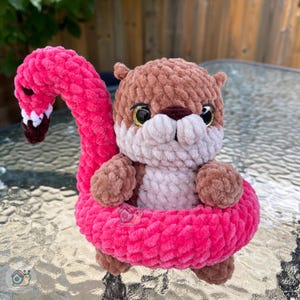 May include: A brown and light beige crocheted otter plush toy sitting inside a bright pink crocheted flamingo-shaped pool float. The otter has large, shiny black eyes and a dark brown nose. The flamingo float features a long, curved neck.