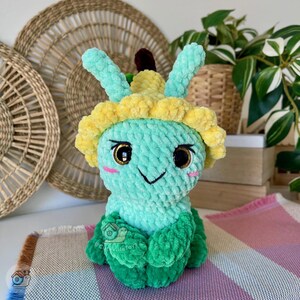 Puede incluir: Un peluche de ganchillo hecho a mano con forma de criatura sonriente de color azul claro con detalles amarillos y verdes. El juguete tiene ojos grandes y expresivos, mejillas rosadas y una sonrisa negra. El peluche est&aacute; sobre una superficie a rayas rosas y blancas.